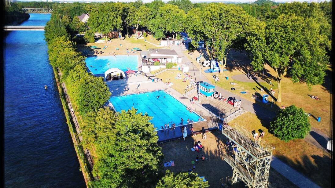 Luftaufnahme eines Schwimmbads mit zwei Becken und Liegewiese, umgeben von Bäumen und einem Fluss im Hintergrund.