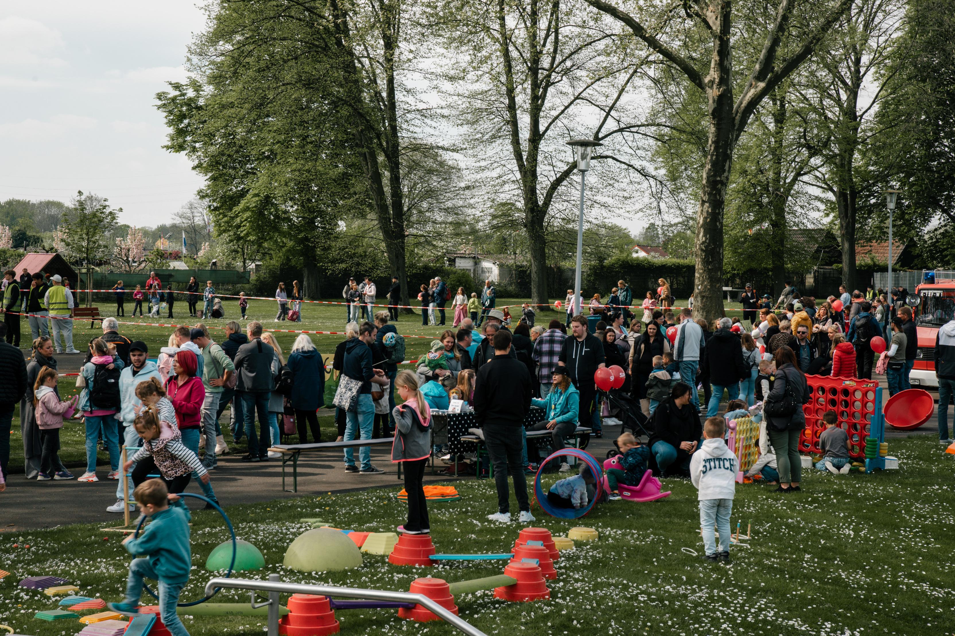 Menschenmenge in einem Park mit Kindern, Spielgeräten und Aktivitäten unter Bäumen an einem sonnigen Tag.