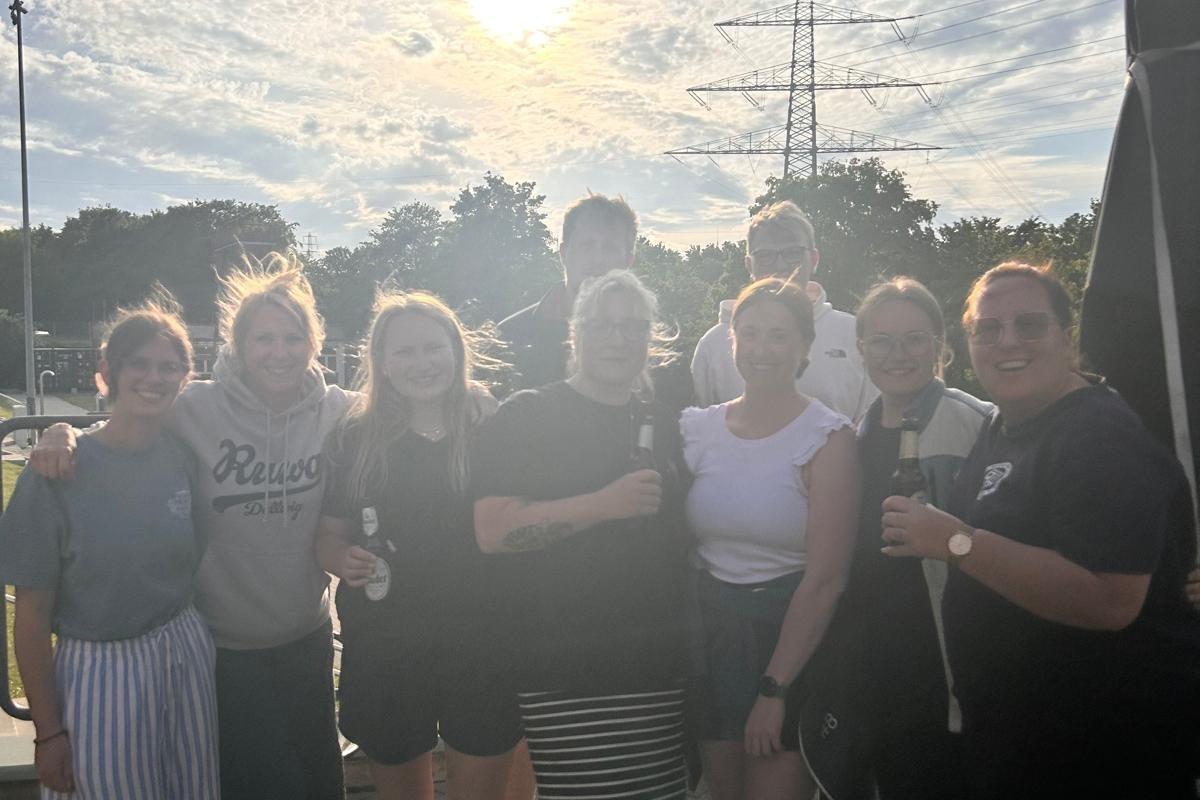 Schwimmlehrer-Gruppe Neun Teilnehmende der Schwimmlehrer-Ausbildung stehen lächelnd zusammen im Außenbereich des Freibads Dellwig. Im Hintergrund sind Bäume, ein Strommast und ein bewölkter Abendhimmel mit Sonnenstrahlen zu sehen. Mehrere Personen tragen
