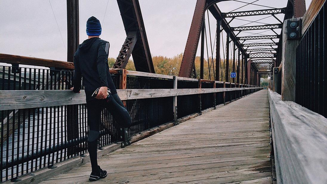 Läufer in sportlicher Kleidung dehnt sich auf einer Holzbrücke mit metallenen Rahmen und herbstlicher Landschaft.