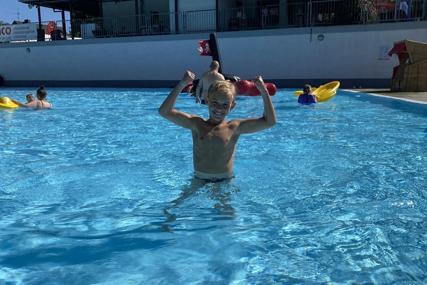 Ein Junge steht im Wasser eines Schwimmbeckens und hebt einen kleinen Bruder auf seinen Schultern.