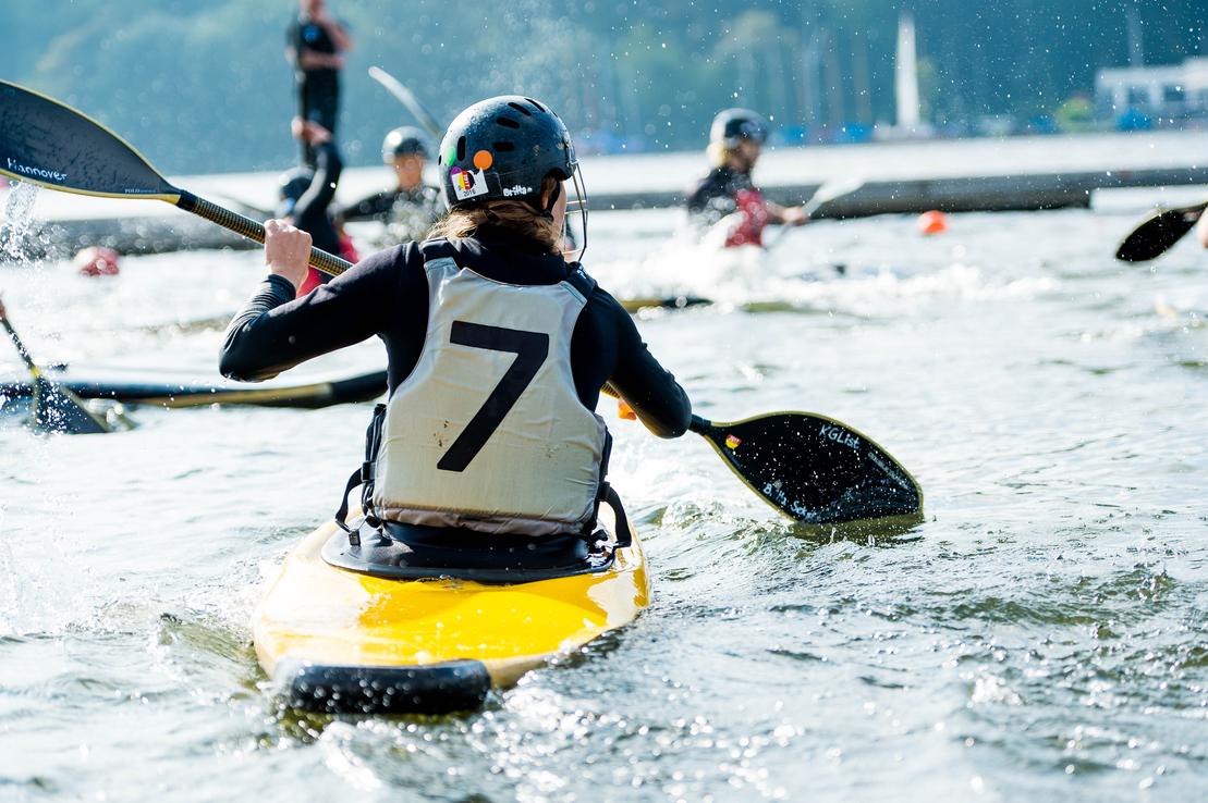 Person im Kanu mit gelbem Boot und paddelndem Arm, auf einem Gewässer mit weiteren Paddlern im Hintergrund.